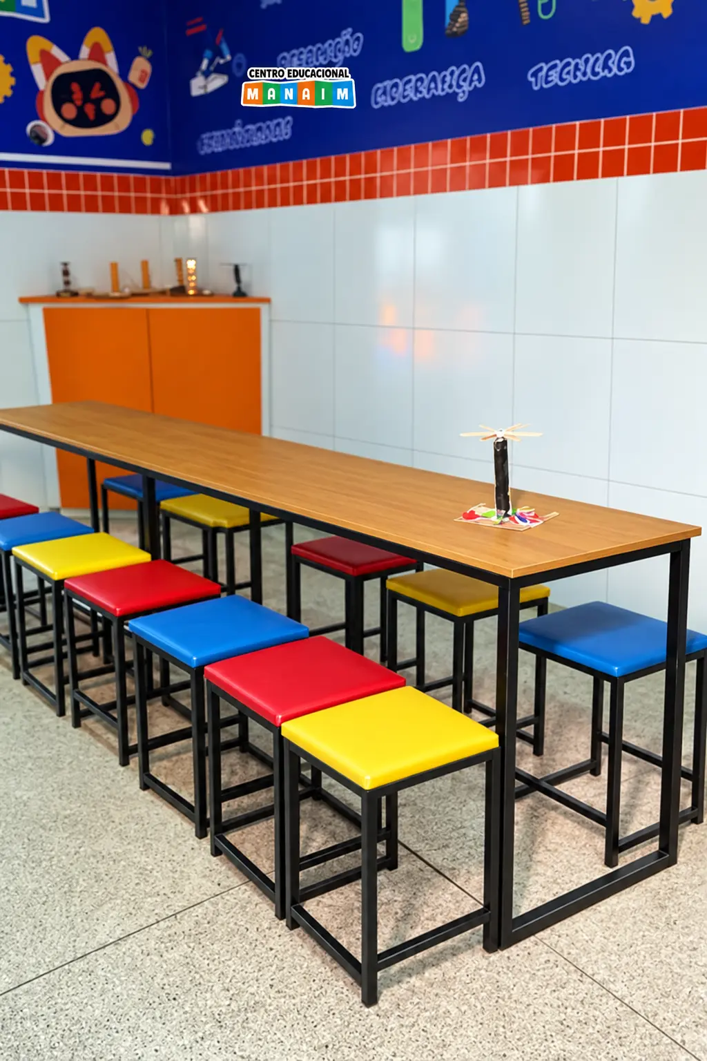 Espaço maker com bancadas de madeira e banquetas coloridas